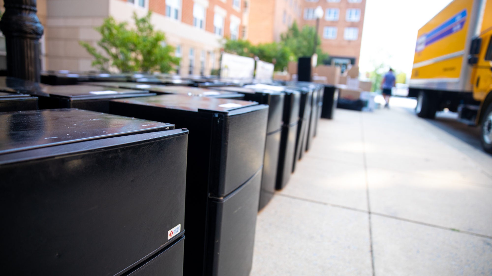 Photo displays a row of mini fridges lined along the sidewalk outside of a dorm.