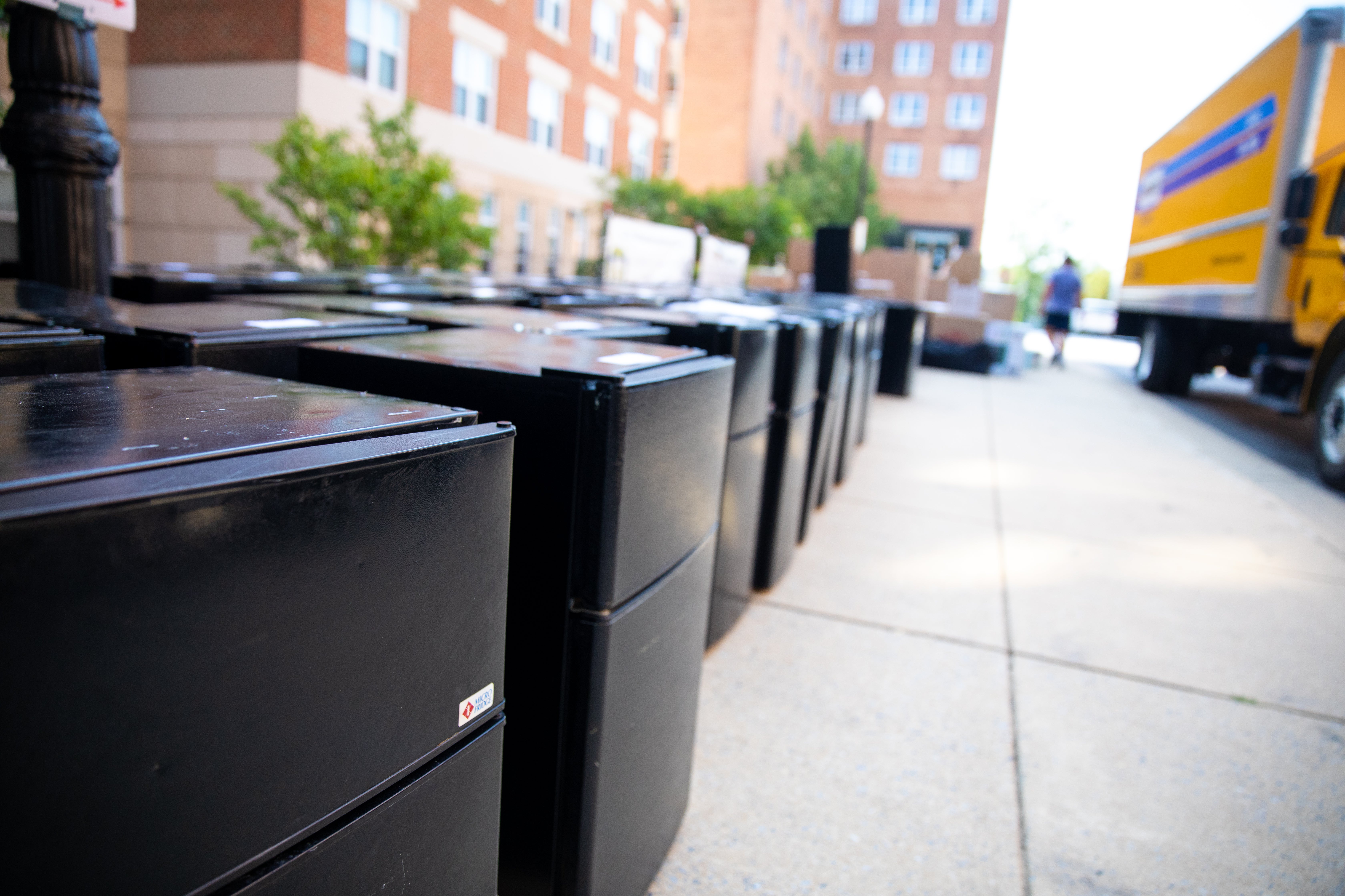 Photo displays a row of mini fridges lined along the sidewalk outside of a dorm.