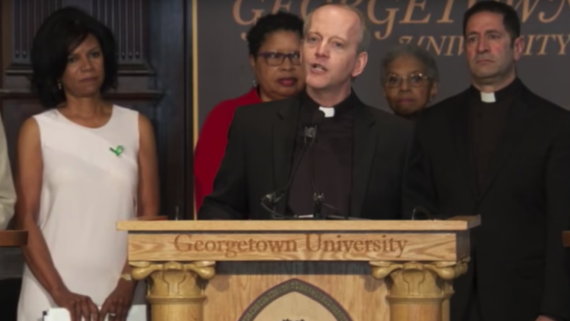 Members of the community descended from people enslaved by the Maryland Jesuits with Jesuits from Georgetown and the Maryland Province with Maryland Province Jesuits and Georgetown Jesuits onstage in Gaston Hall with Jesuit speaking at podium