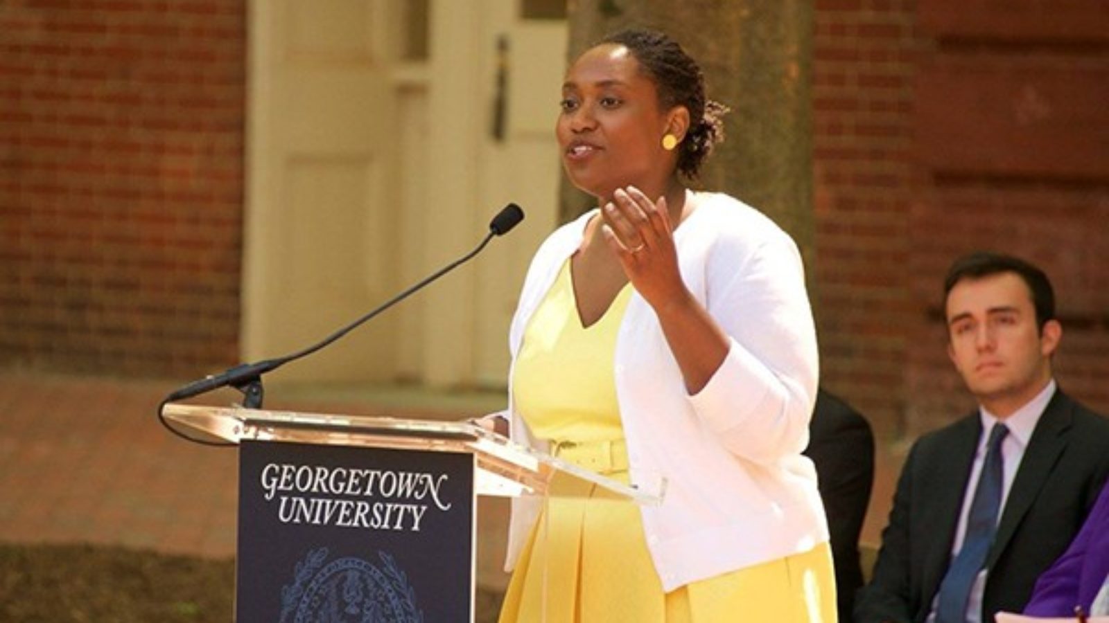 Marcia Chatelain speaking into a microphone with man sitting and looking toward her