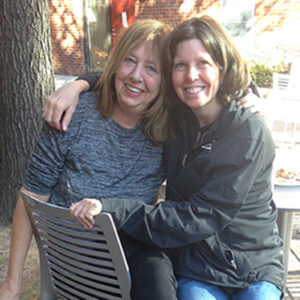 Elizabeth Velez and Mary Esselman sit on a chair outside with Mary's arm around Elizabeth