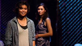 Cristina Ibarra speaks on a theater stage with another actress behind her.