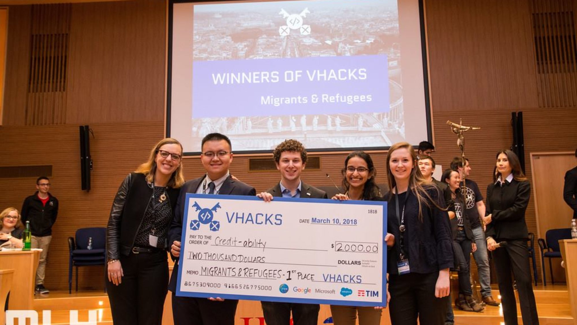 Roisin McLoughlin, Jake Glass, Lucy Obus, Rushika Shekhar and Yanchen Wang stand on a stage holding an oversized check address to Credit-ability for $2,000 with other people and a screen behind them reading Winners of Hacks, Migrants and Refugees