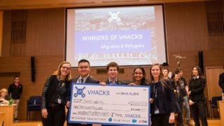 Roisin McLoughlin, Jake Glass, Lucy Obus, Rushika Shekhar and Yanchen Wang stand on a stage holding an oversized check address to Credit-ability for $2,000 with other people and a screen behind them reading Winners of Hacks, Migrants and Refugees