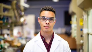 Orlando Stewart stands wearing a white lab coat and goggles in a laboratory.