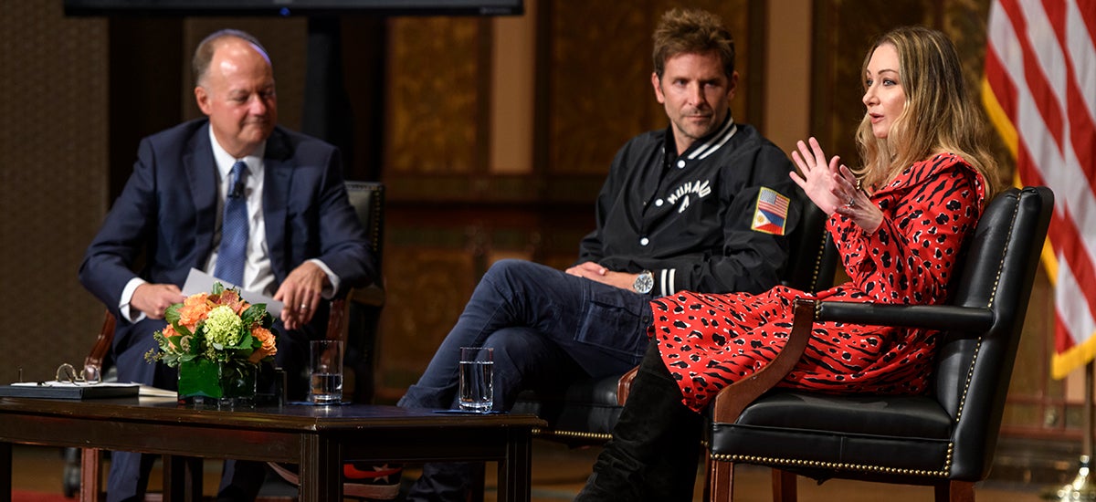 Bradley Cooper looks on as Blair Rich speaks to the audience as they are seated on stage.