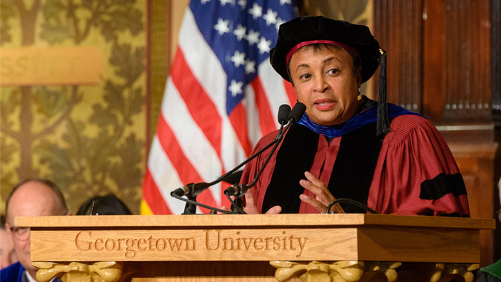 Carla Hayden at Georgetown University podium wearing cap and gown with American flag behind her