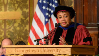 Carla Hayden at Georgetown University podium wearing cap and gown with American flag behind her