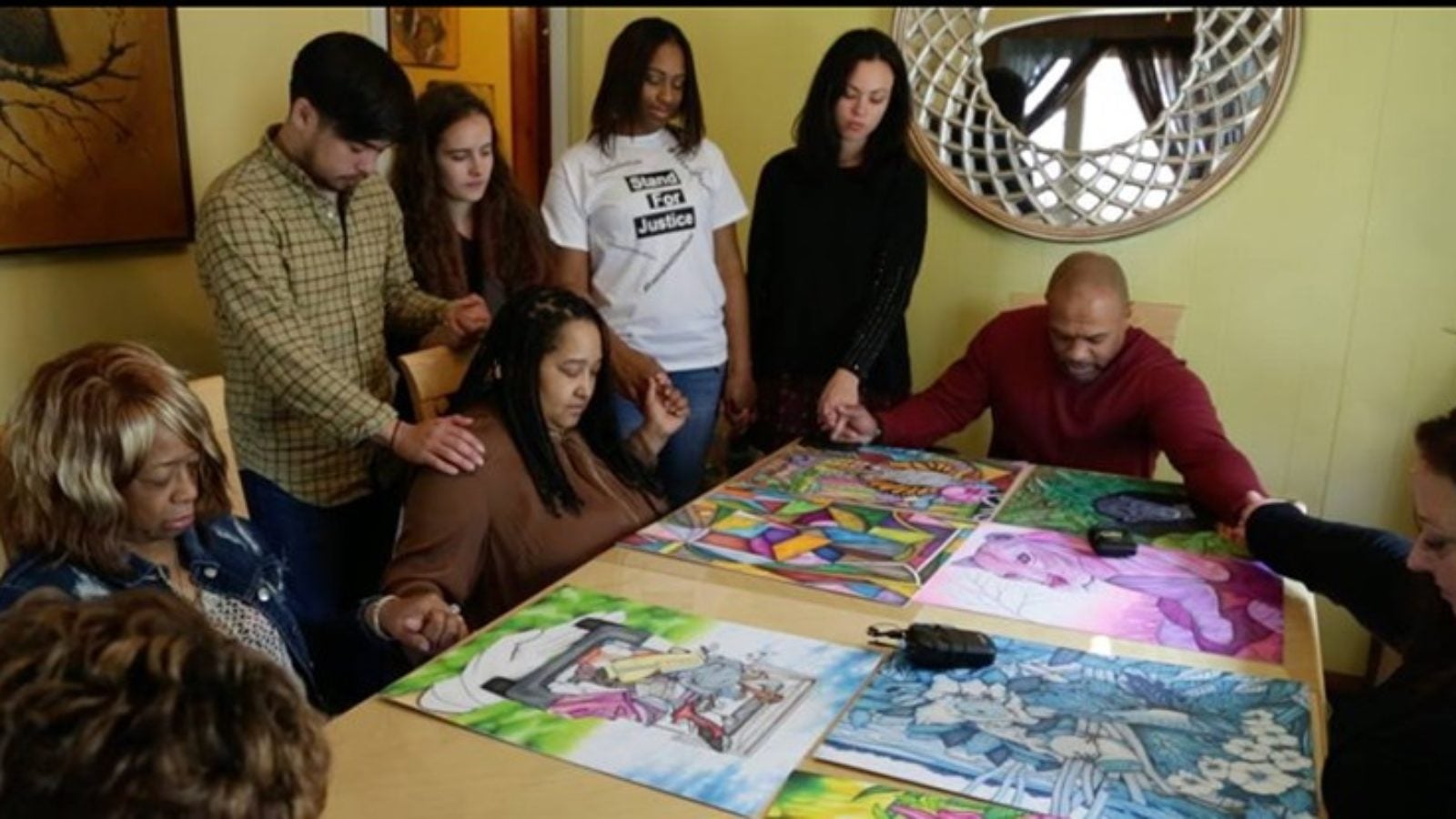 Valentino Dixon and family and Georgetown students hold hands and pray around table with Valentino Dixon&#039;s artwork