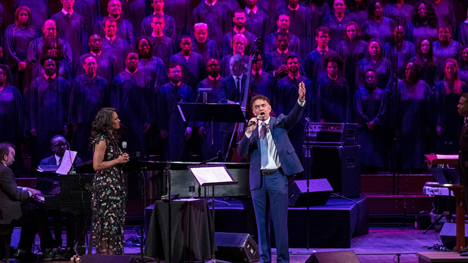 Audra McDonald looks on as Brian Stokes Mitchell sings with a choir singing on stage with him in the background.