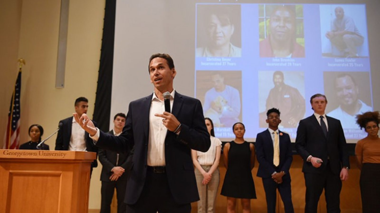 Marc Howard standing with microphone in front of students and video screen of six incarcerated individuals