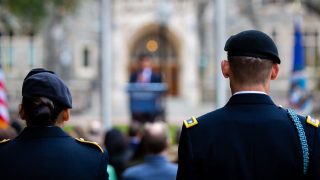 A female and male member of the military stand in uniform facing White-Gravenor Hall with an American flag in the foreground.