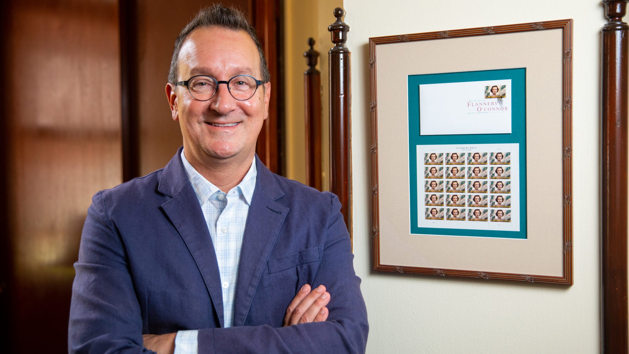 Mark Bosco stands with arms folded next to frame with Flannery O&#039;Connor postage stamp