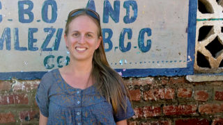 Sara Fischer stands outside in front of a sign hanging on a brick building.