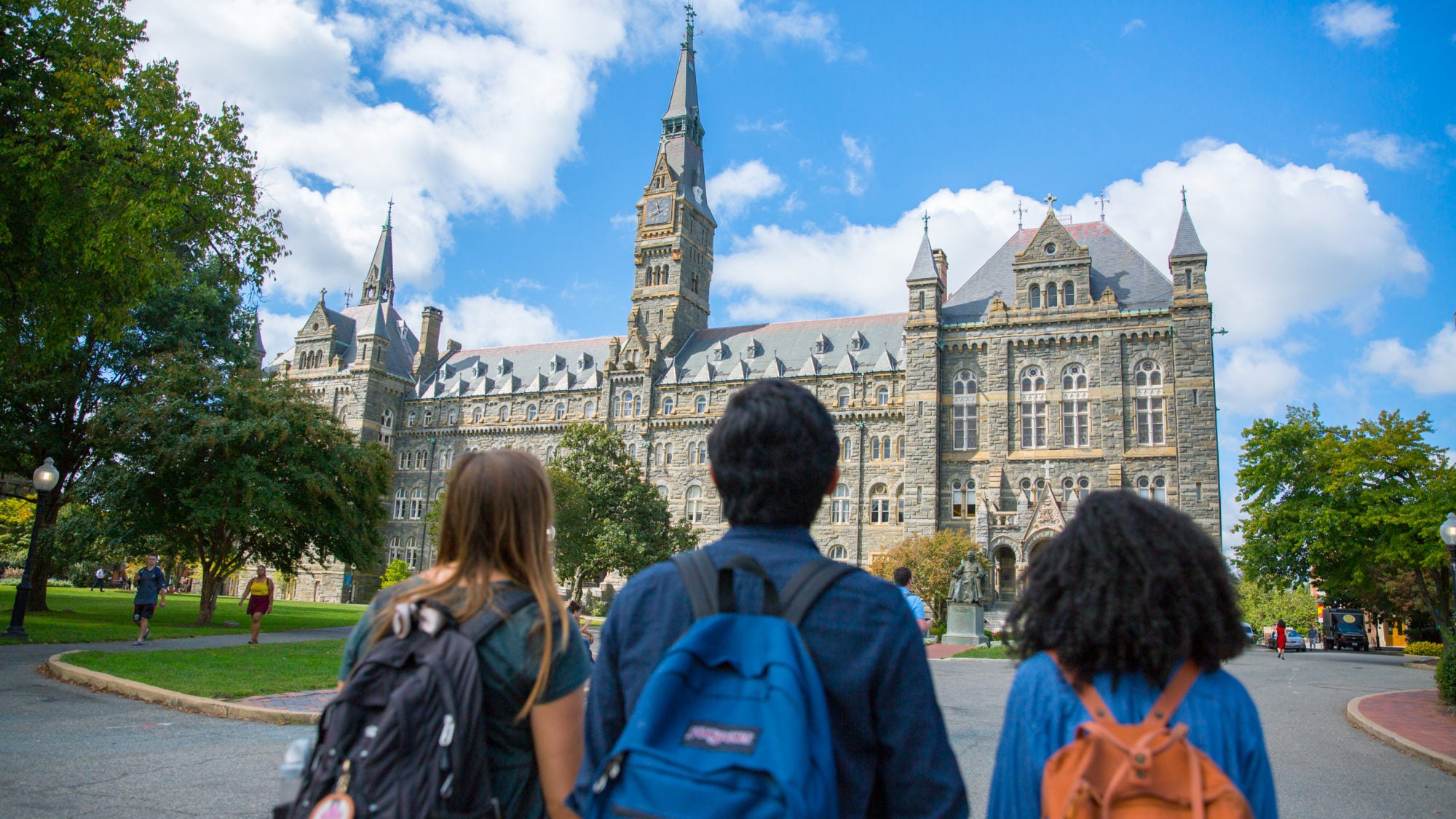 Three undergraduate students are walking towards Healy Hall. The backs of their heads and backpacks are visible as they walk towards the building.