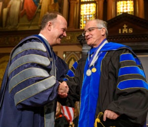 John J. DeGioia shakes the hand of Italo Mocchetti on stage while they are both dressed in academic regalia.