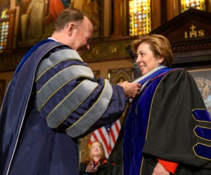 John J. DeGioia shakes the hand of Cristina Sanz on stage while they are both dressed in academic regalia.