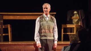 David Strathairn on stage with a desk and chairs playing Jan Karski