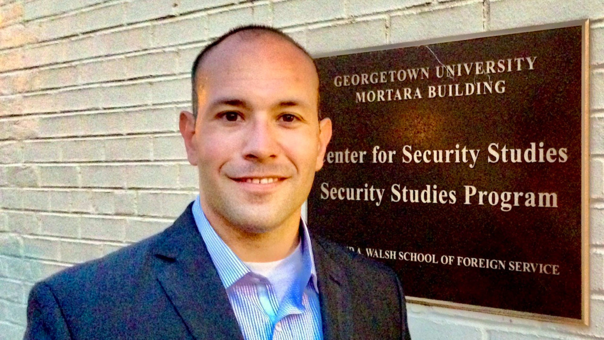 Chris Mercado stands near the Peace and Security Studies building plaque.