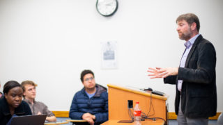 Adam Rothman gestures standing near a podium in a classroom with three students listening