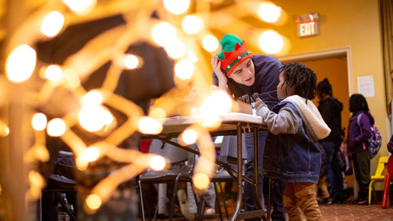 A student wearing a Christmas hat leans down to talk to a child with decorative lights in the foreground.