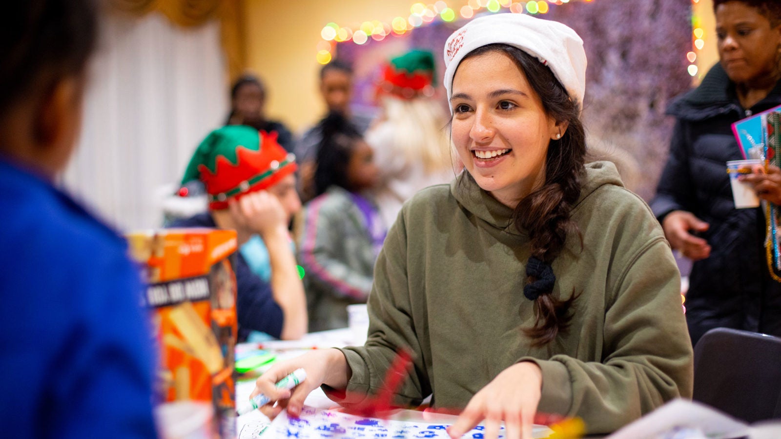 A female student wears a Santa hat while sitting down and talking to a young child.