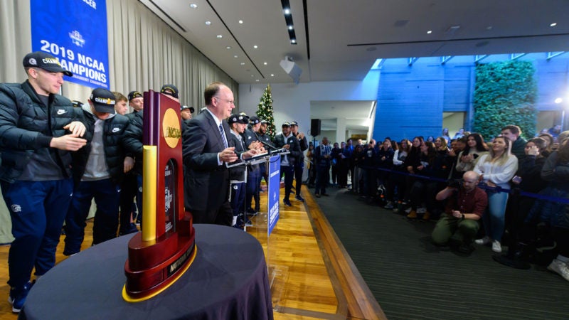 Members of teh soccer team gather on stage as John J. DeGioa speaks to a crowd of cheering students with the championship trophy in the foreground.