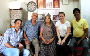  Kyra Kocis sits with a family in India with stacks of cloth and photos and art behind them.