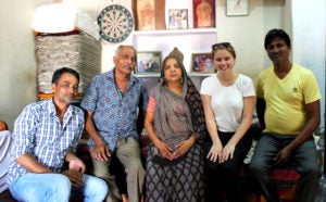 Kyra Kocis sits with a family in India with stacks of cloth and photos and art behind them.