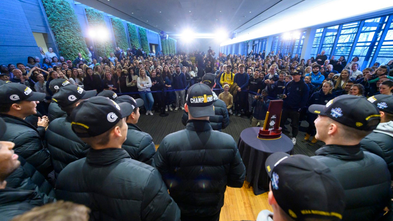 Members of the Georgetown soccer team are welcomed by a crowd of Georgetown students in a room with plants growing on the walls