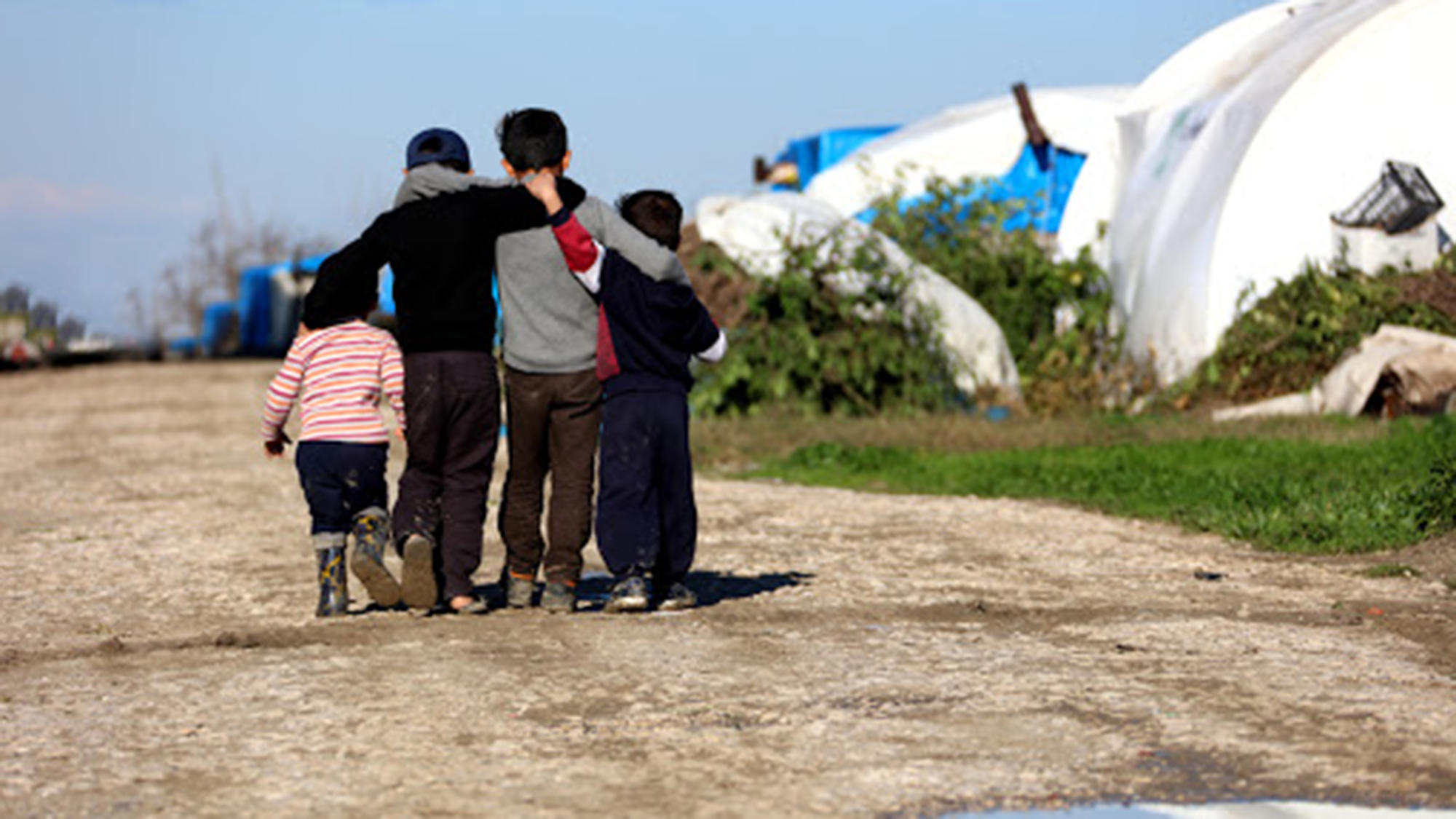 Four refugee children walking on a dirt road near tents