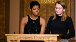 Brianna Rodgers and Lucy Chatfield stand beside one another on stage and speak at a lectern to the audience.