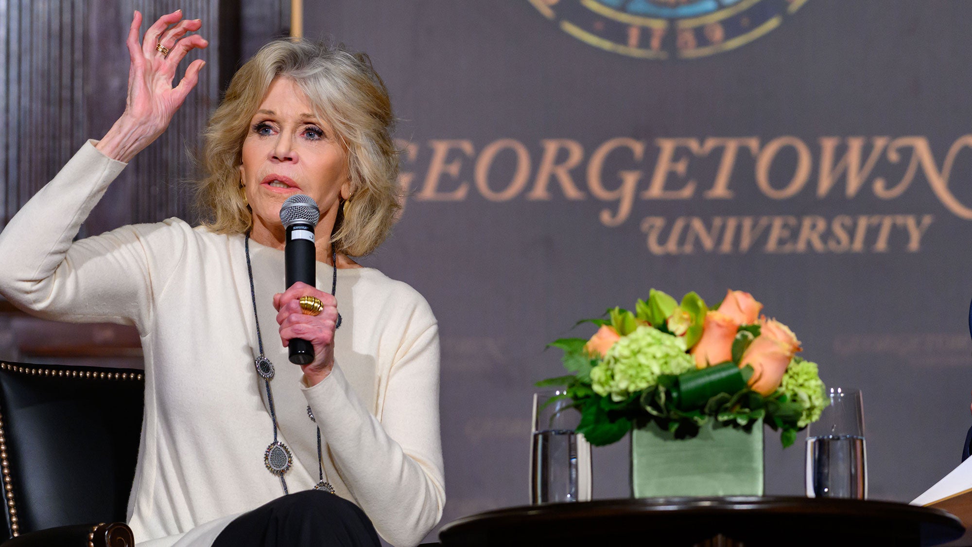 Jane Fonda raises her hand while sitting on stage.