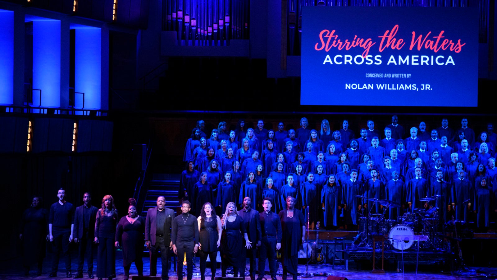 Singers dressed in black on a darkly lit stage perforrm with a standing choit in the background and a screen reading &quot;Stirring the Waters.&quot;