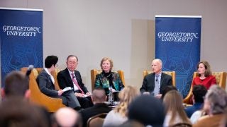 Desha Girod holds a microphone sitting next to Ban Ki-Moon, Mary Robinson, Jerry Brown and Rachel Bronson with a Georgetown University banner behind them