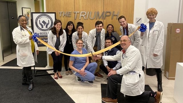 DDr. Stephen Ray Mitchell kneels on the floor cutting a yellow ribbon held by two other doctors as doctors and other people kneel or squat in front of a sign reading The Triumph