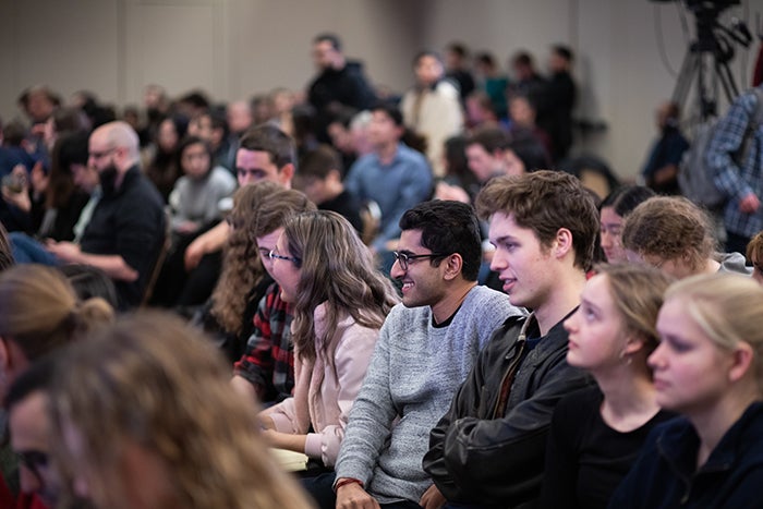 An audience of mostly students sits and stands in a packed room