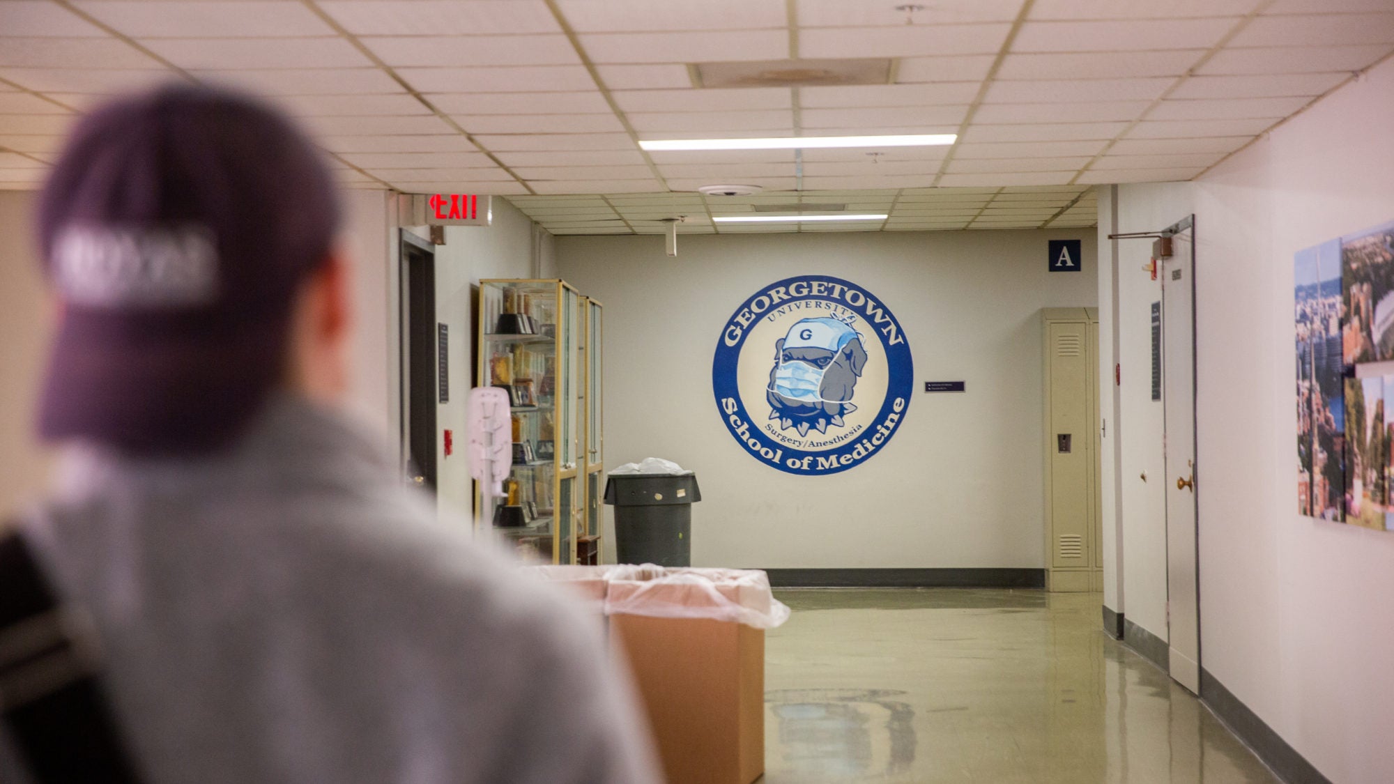 A student with a blue hat on backwards walks down a hallway, at the end of which a logo for the School of Medicine is painted on the wall.