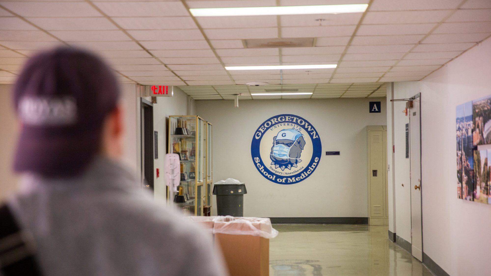 A student with a blue hat on backwards walks down a hallway, at the end of which a logo for the School of Medicine is painted on the wall.