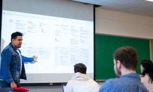 Juan Manuel Menjîvar stands in front of class and teaches his lessons projected on a white screen while studetns look on.