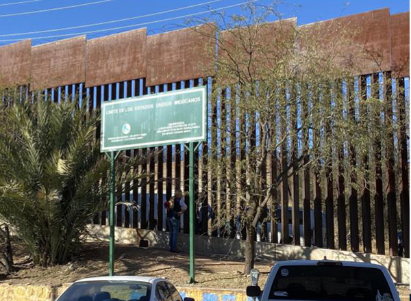 A person talks through the wall at the border of the United States and Mexico with a sign in front of it and cars.