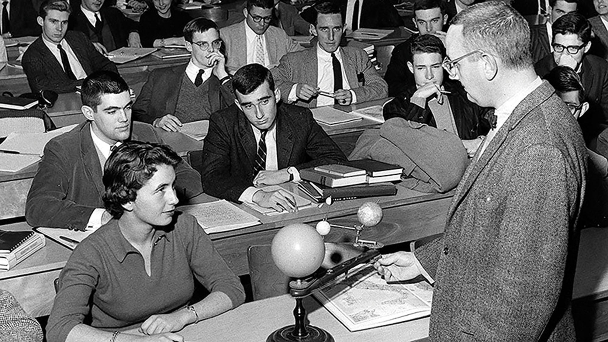 A woman sits in a classroom surrounded by men while the male professor teaches.