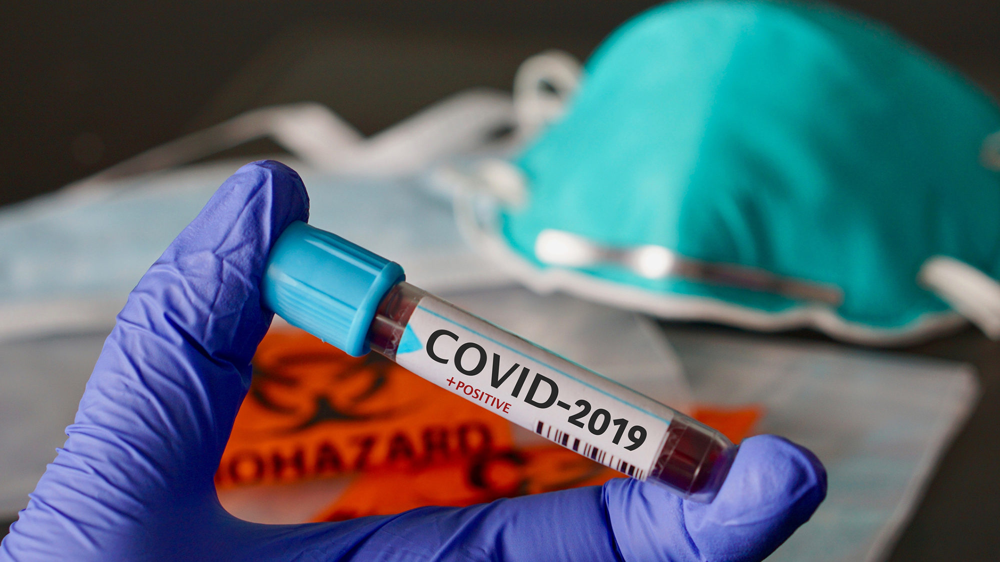 a mask lying on a table with a bag labeled biohazard and a gloved hand holding a test tube filled with blood with the words COVID-19 on it.