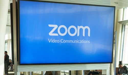 A screen with Zoom video communications on it with people milling around it. 