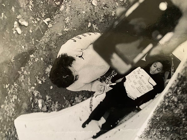 Photo of a hand taking an iPhone photo of a woman lying down wearing a sign on her chest reading "The System Was Never Broken It was BUILT this way" and another woman crouched over her.