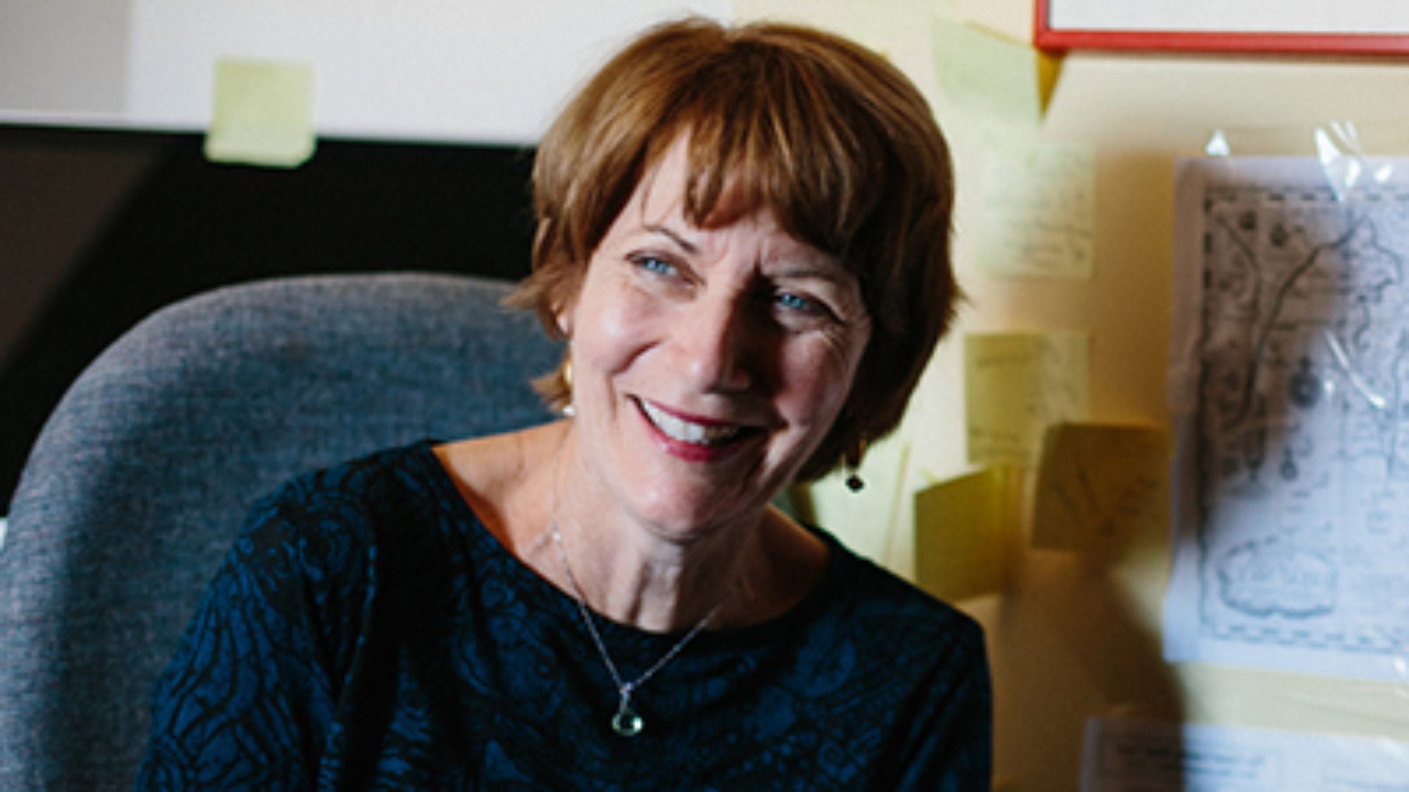 Maureen Corrigan sitting in a chair with post-it notes and a map on the wall behind her