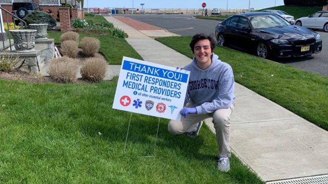 Liam Marshall squatting on a front lawn with a sign stuck in the lawn reading &quot;Thank You First Responders and Medical Providers and all other essential workers.&quot;