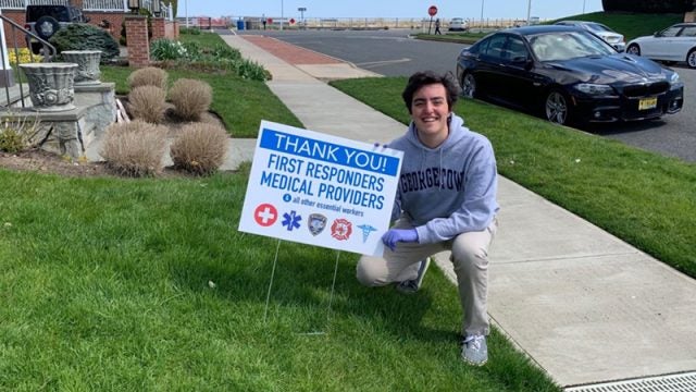 Liam Marshall squatting on a front lawn with a sign stuck in the lawn reading &quot;Thank You First Responders and Medical Providers and all other essential workers.&quot;