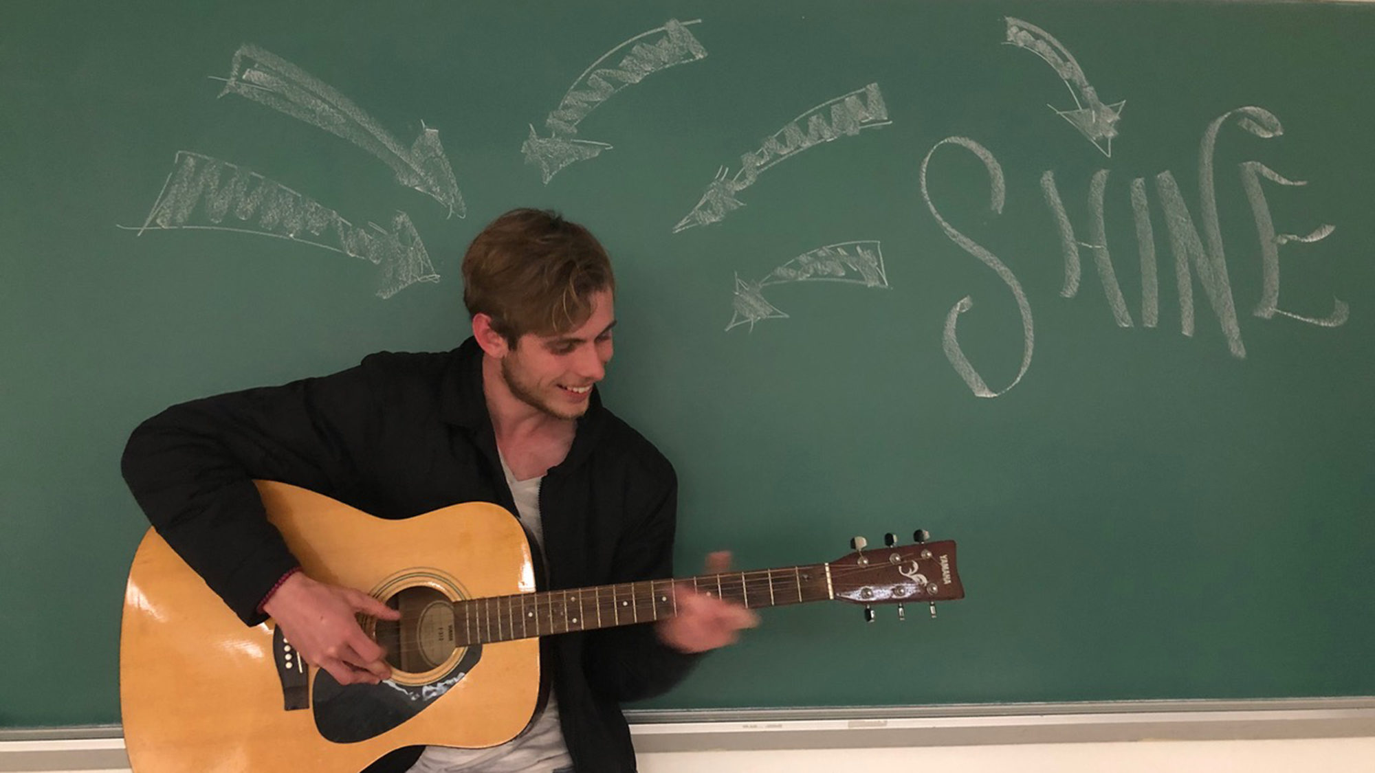 Kent Hall playing guitar in front of a black board chalked with arrows pointing toward him and another arrow pointing away from him and the word Shine.