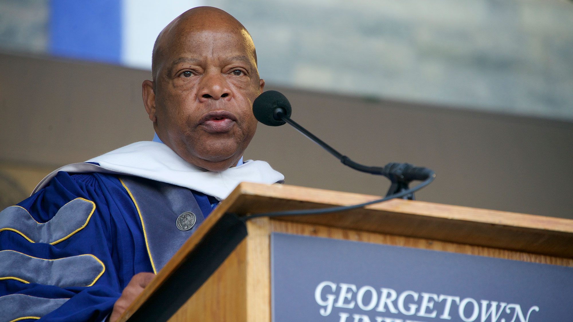 John Lewis stands at a lectern speaking into a microphone while wearing academic regalia.